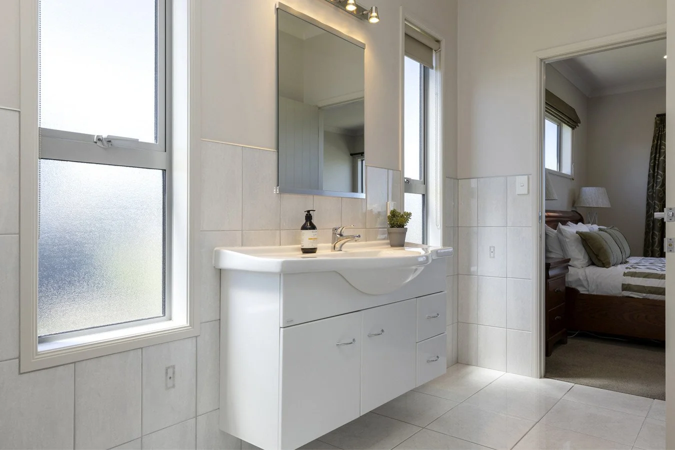 Bathroom with white sink and cabinet, large mirror, two windows, and bedroom visible through open door.