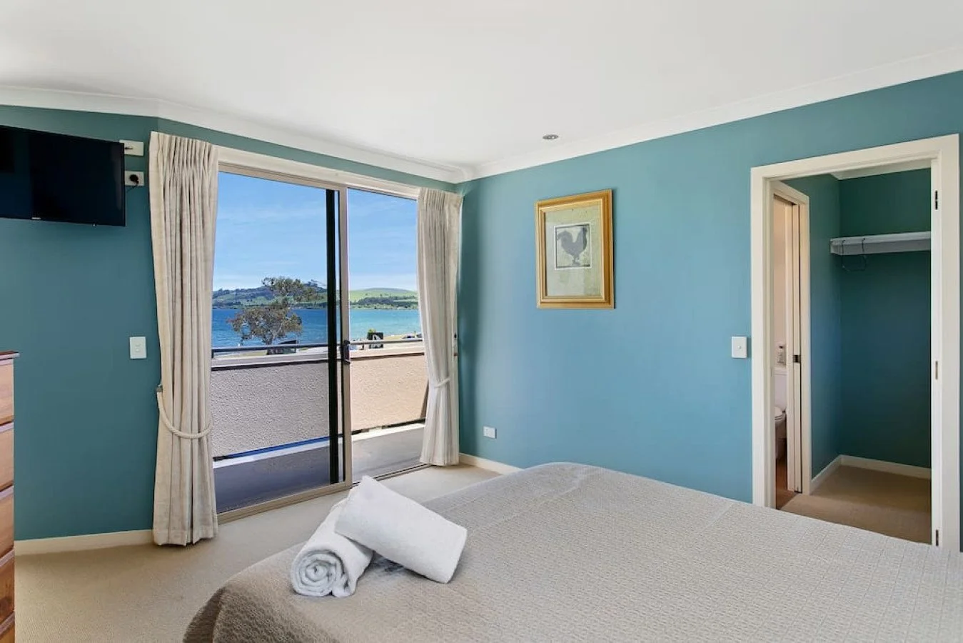 Bedroom with sliding glass door opening to balcony view of water and greenery, beige bed with rolled towels, sea-themed artwork, TV, and closet.