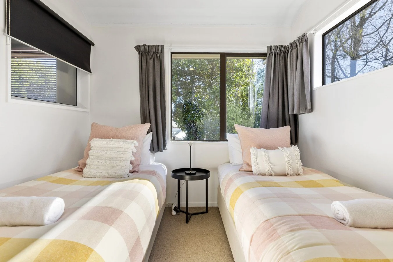 A bedroom with two twin beds, each with pastel-colored checkered comforters and pillows, a black side table with a lamp between the beds, and windows with grey curtains allowing natural light to enter.