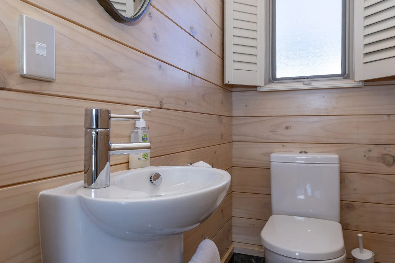 Small bathroom with wooden panel walls, a white toilet, a small white sink with a chrome faucet, a soap dispenser, a mirror, and a window with white shutters.