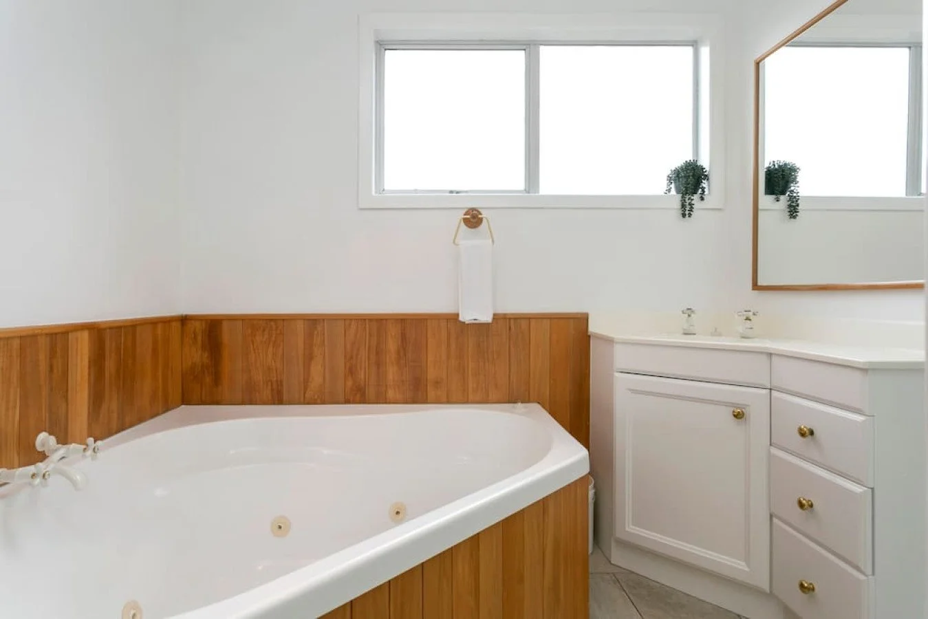 Bathroom with white walls, wooden paneling around a bathtub, a small white cabinet with gold knobs, a large mirror, and two small potted plants on the window sill.