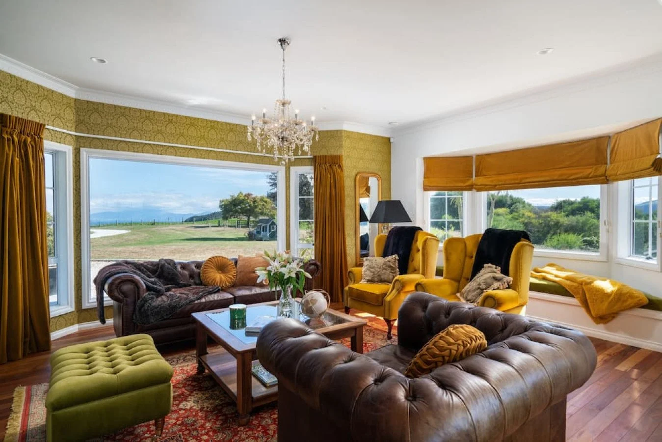 Living room with yellow and brown furniture, large windows showing a green outdoor landscape, chandelier, floral arrangement on coffee table, and patterned rug.