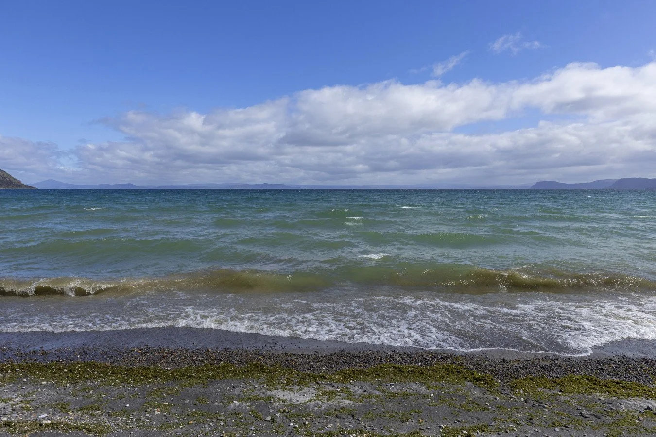 A view of a sandy beach with small waves, green seaweed, and pebbles, under a partly cloudy blue sky with distant islands on the horizon.