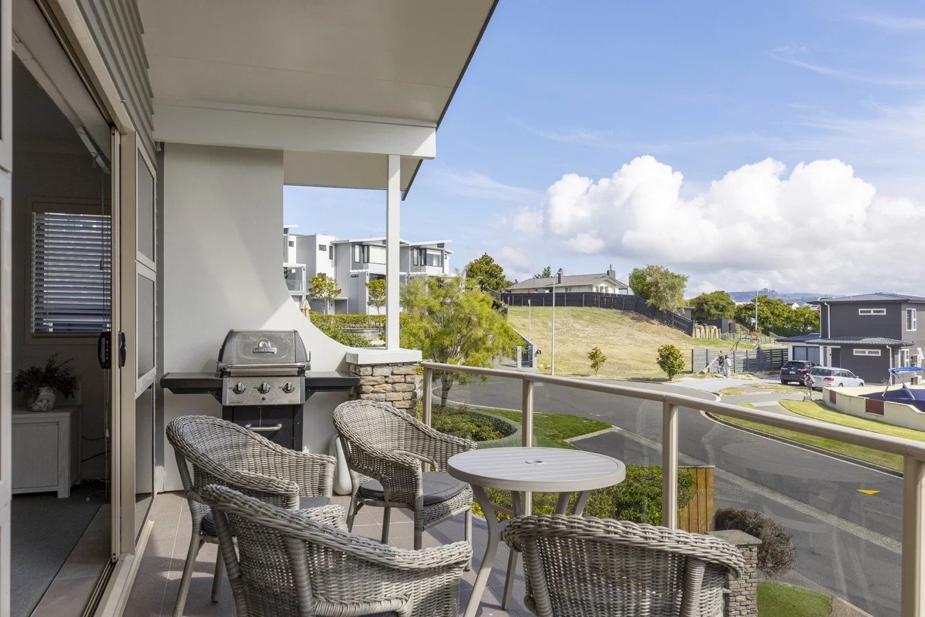 Balcony with wicker chairs and a small round table, view of suburban neighborhood with houses, grassy hill, and blue sky with clouds.