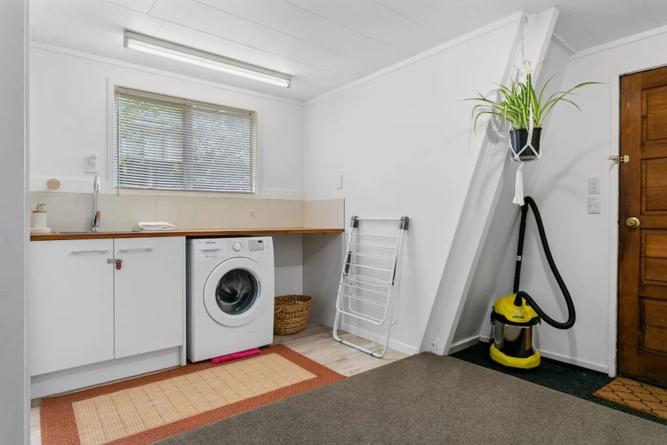 Laundry room with washing machine, laundry drying rack, vacuum cleaner, window with blinds, potted plant, and wooden door.