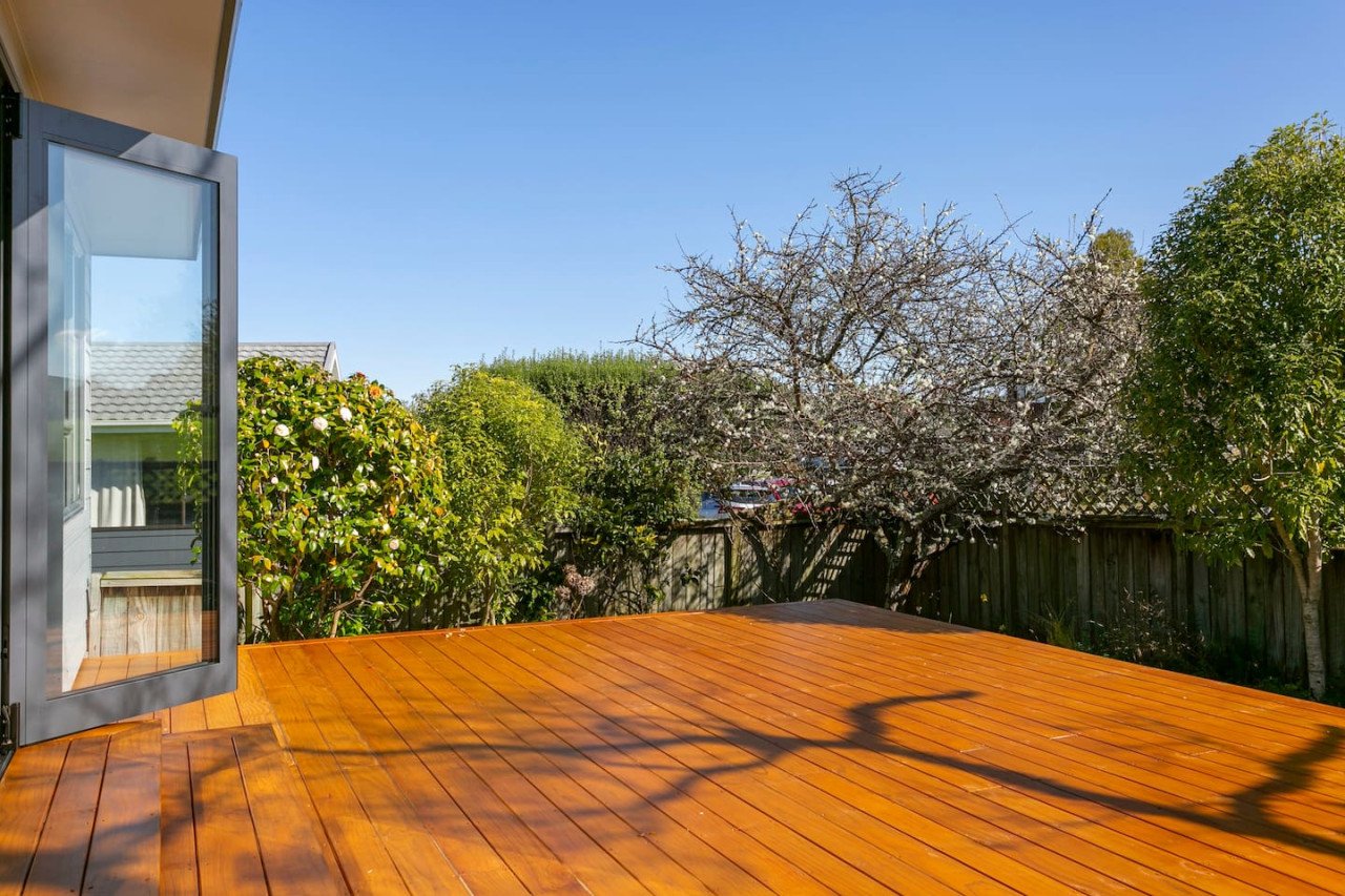 Open window on a house overlooking a wooden deck, with trees and bushes, one tree with blossoms, in a sunny outdoor setting.