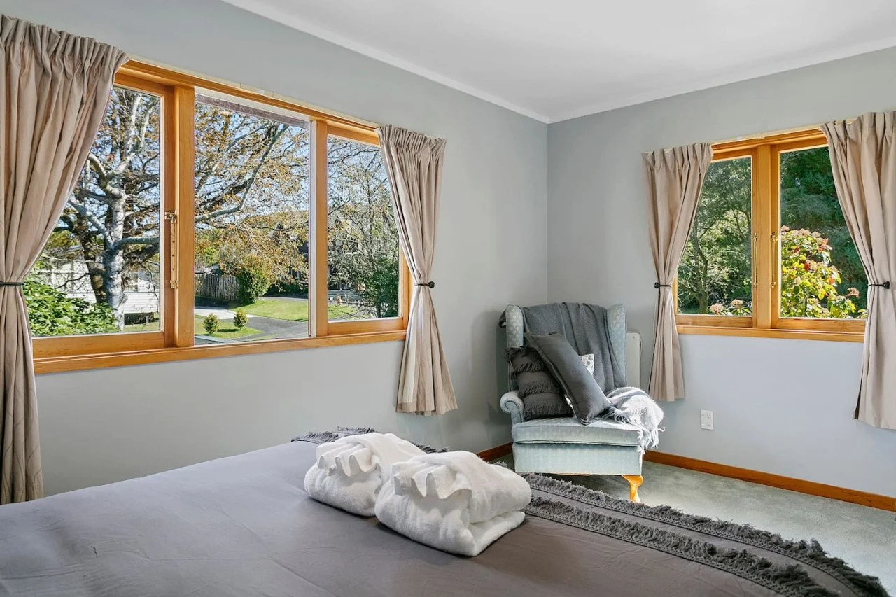 Bedroom with two large windows, beige curtains, a bed with folded towels, an armchair with pillows and a throw, and a view of trees outside