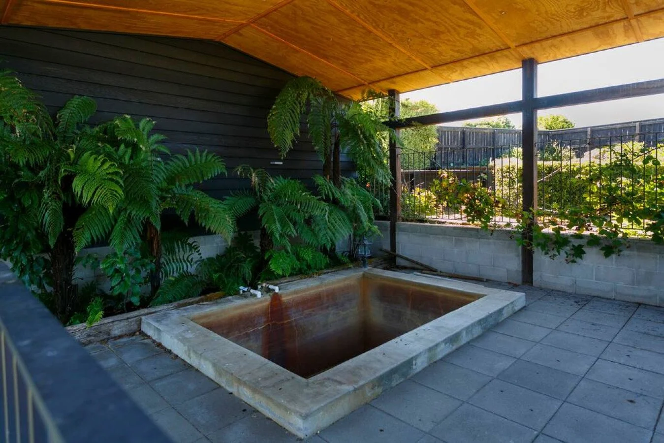 Outdoor covered patio with a small rectangular heated or cooled pool, surrounded by grey tiles, lush green plants, a black wooden house wall, and a wooden ceiling. The patio is enclosed by a black metal fence with greenery outside.