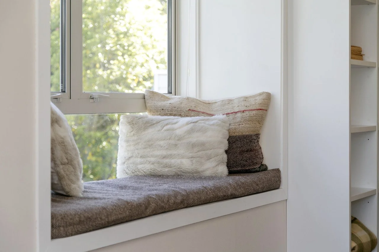A cozy window nook with a cushioned seat and decorative pillows, overlooking greenery outside.