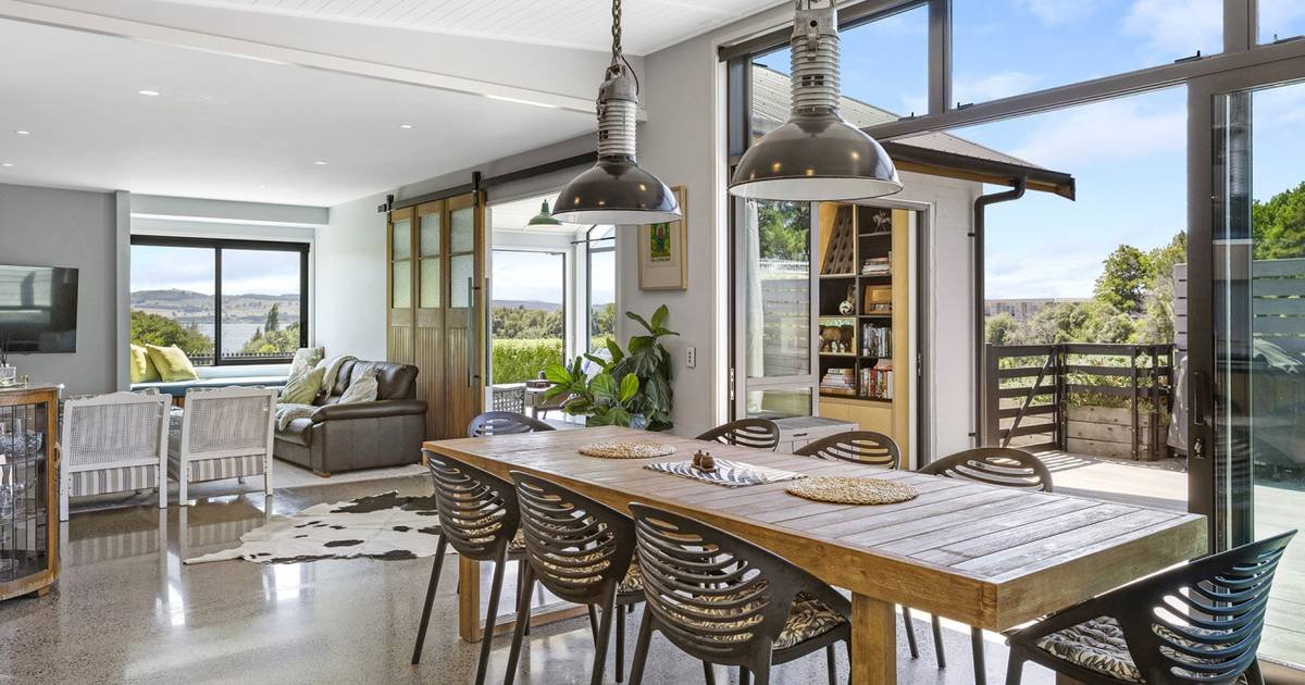 Open-concept living and dining area with large windows, wooden dining table with black chairs, sliding glass door leading to an outdoor balcony, and a view of greenery and water in the background.