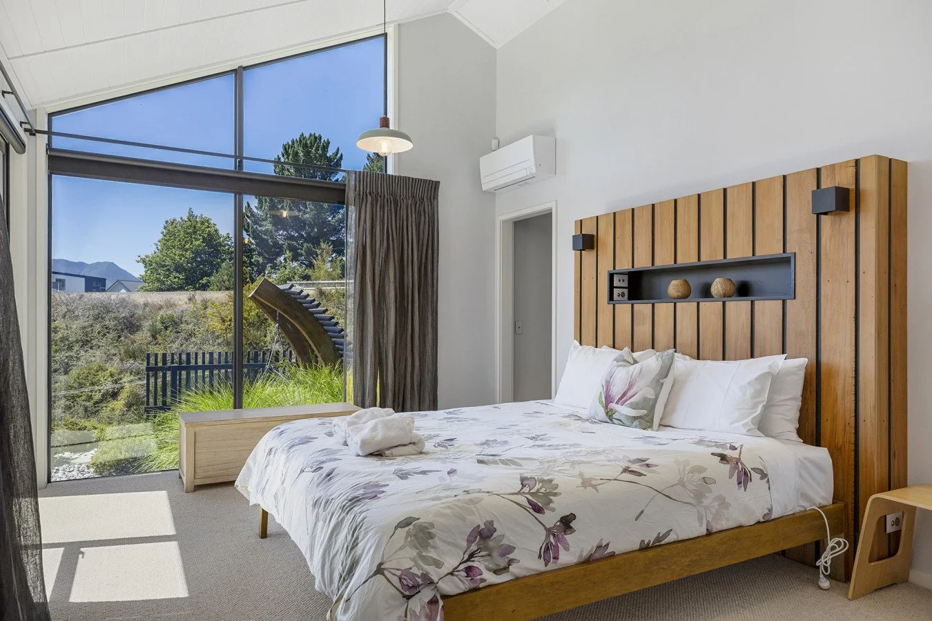 A bedroom with a large window showing a scenic outdoor view, a bed with floral bedding, and a wooden headboard with built-in shelf decorations.
