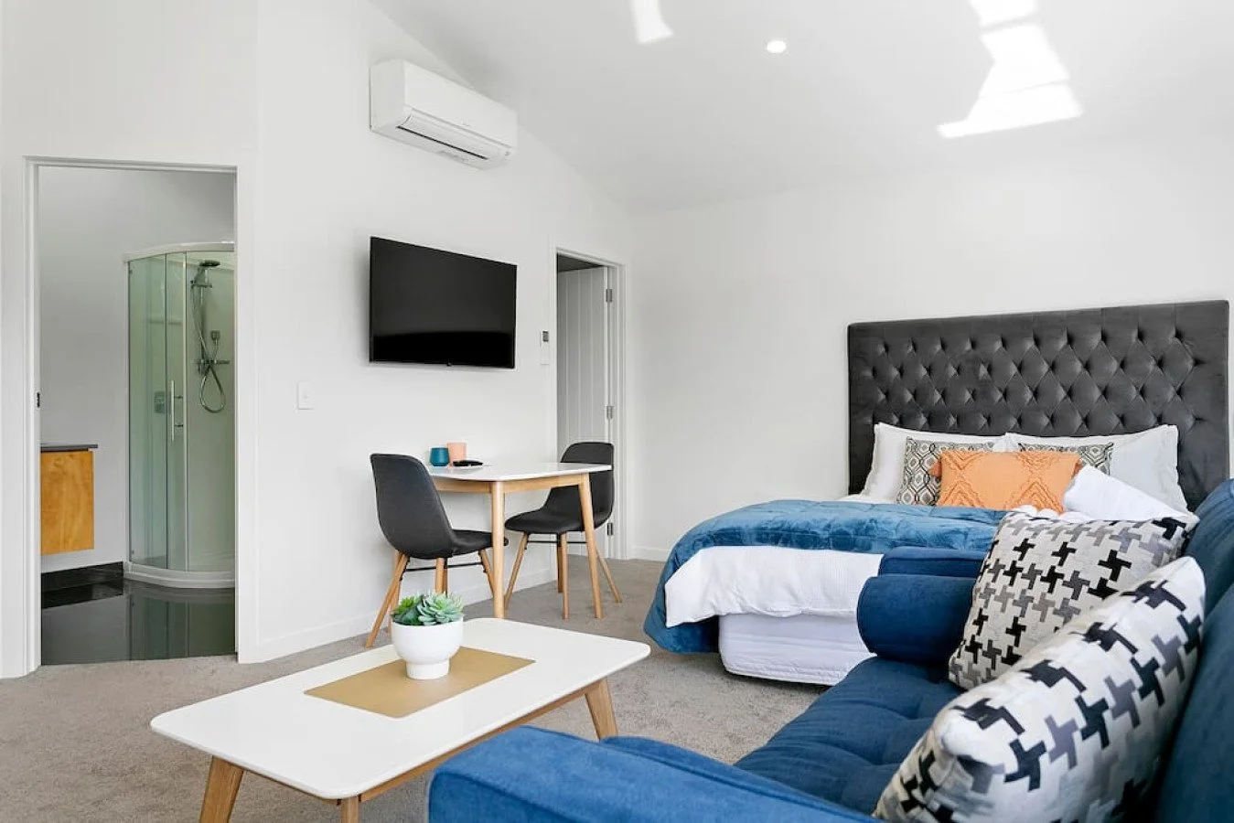 Modern hotel room with a bed, blue sofa with patterned pillows, a small white coffee table with a plant, wall-mounted TV, dining table with two chairs, and a visible bathroom with a shower.