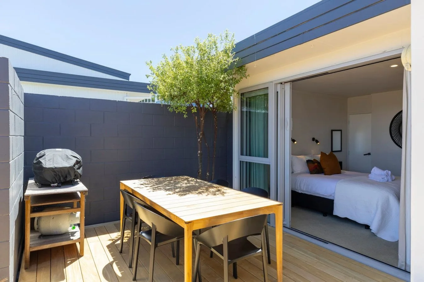 Outdoor patio with wooden dining table, black chairs, a barbecue covered with a black cover, and a small tree next to a dark grey wall, adjacent to a bedroom with sliding glass door.