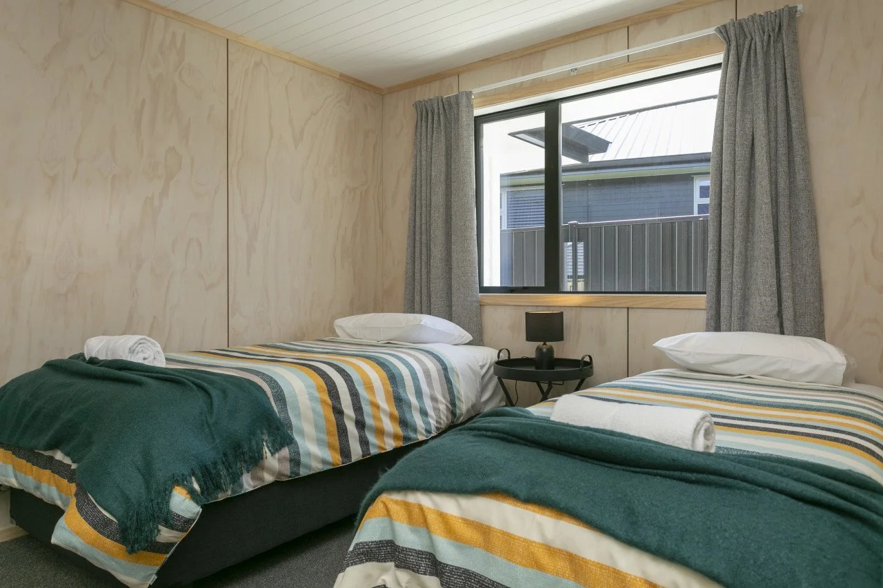 A bedroom with two twin beds with striped bed covers, pillows, and rolled towels, a side table with a black lamp, a window with gray curtains, and exterior view of a fence and neighboring house.