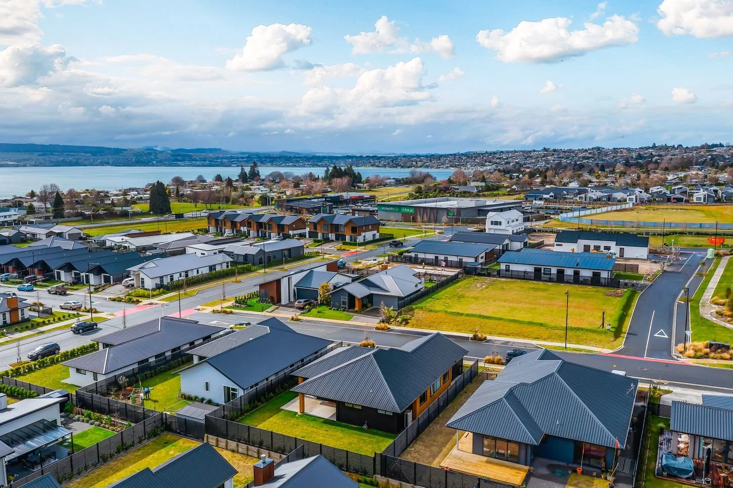 Aerial view of a suburban neighborhood near a large body of water, with modern houses, green lawns, roads, and a cloudy sky.