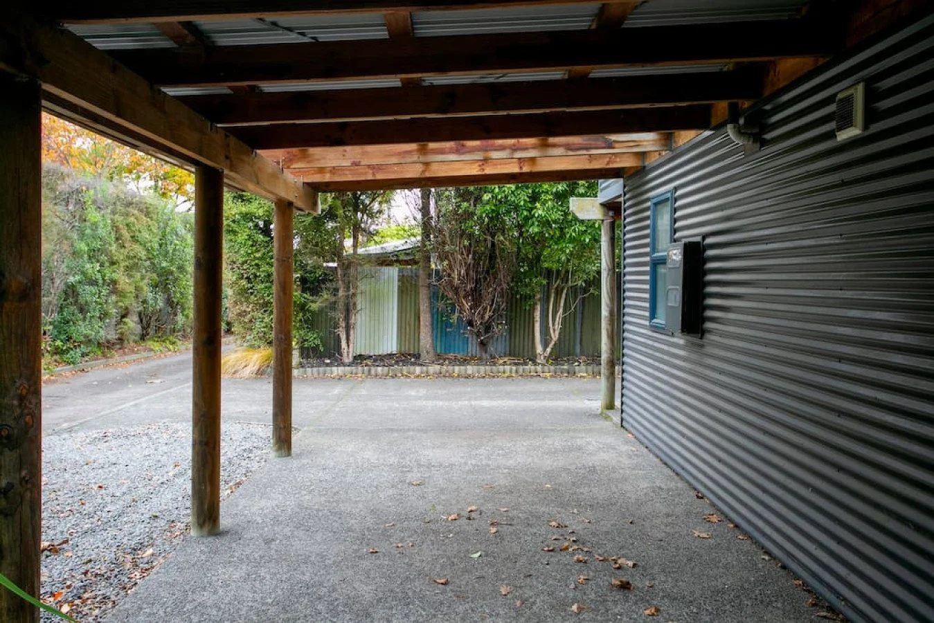 Covered carport with wooden beams and gravel driveway, surrounded by trees and a metal fence.