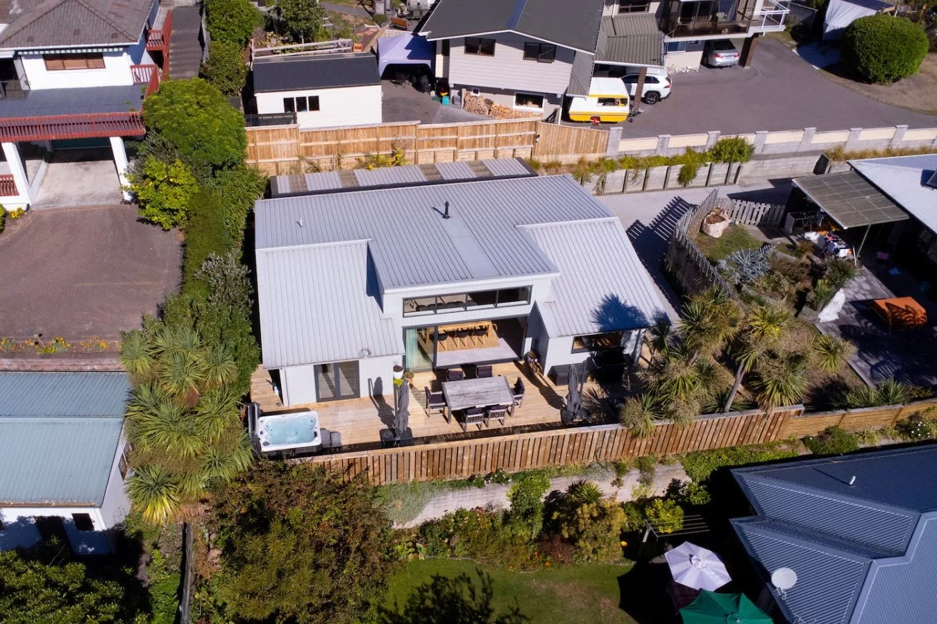 Aerial view of a modern house with a metal roof, outdoor patio with dining table and hot tub, surrounded by trees and other residential buildings.