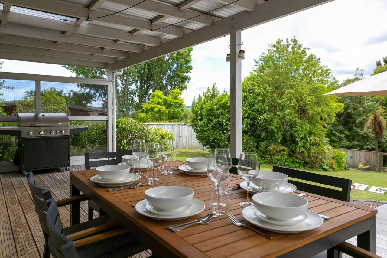Outdoor patio with a wooden dining table set with white bowls, plates, and glassware, on a deck with greenery and trees in the background, and a barbecue grill on the side.