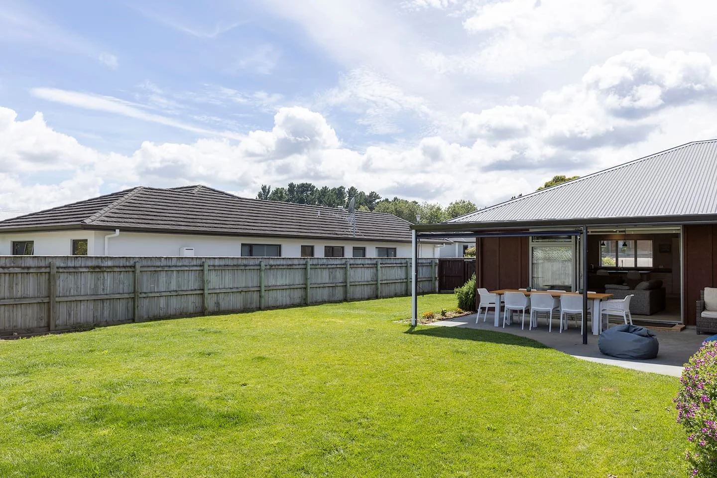 Backyard with green lawn, wooden fence, house with gray roof, patio with outdoor dining table and chairs, bean bag, and garden with a flowering shrub under a partly cloudy sky.