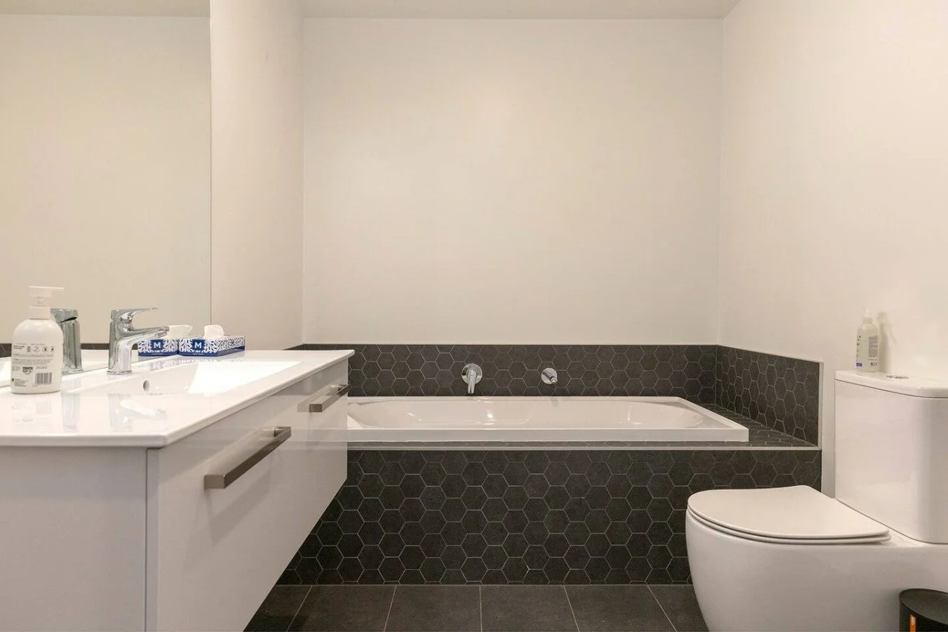 Modern bathroom with white vanity, faucet, soap dispenser, bathtub with black hexagon tile surround, and white toilet.