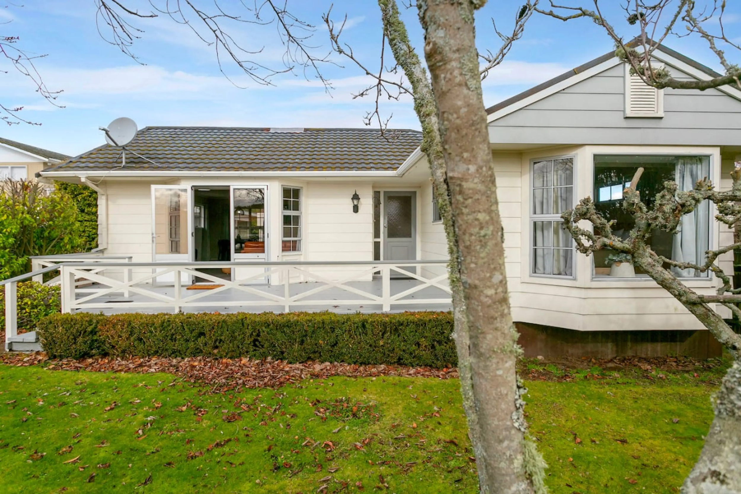 View of a white house with a gray roof, a front door, large windows, a small porch with a white railing, and a green lawn with scattered leaves in the foreground. A tree with moss on its branches partially obscures the right side of the house.