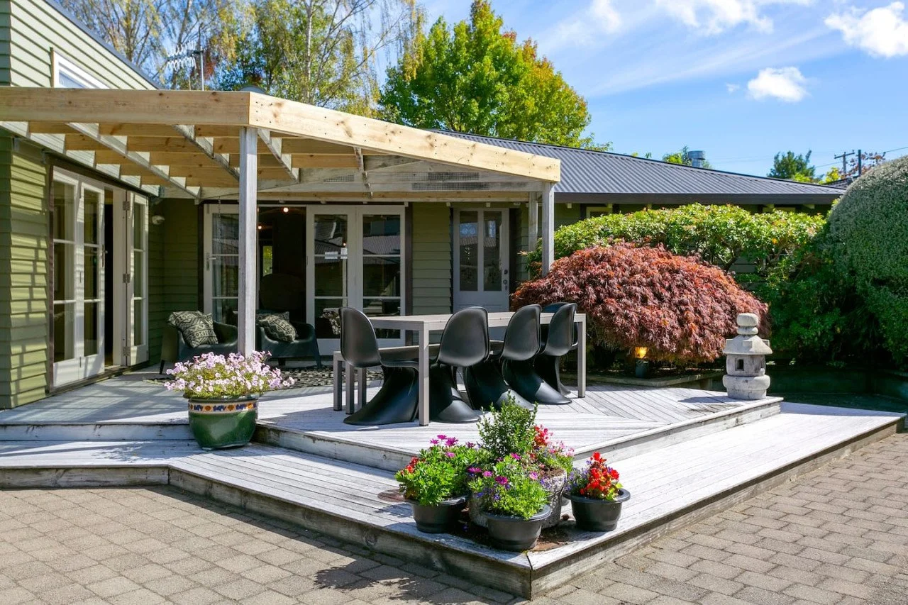 A backyard patio with a wooden deck, black outdoor dining chairs around a table, potted flowers, and greenery including trees and shrubs, with a house in the background and a bright blue sky.