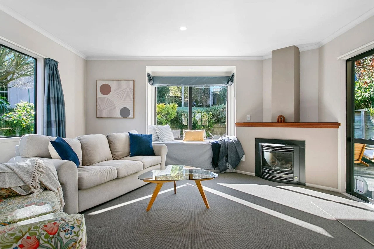 Bright living room with beige sofa, blue pillows, floral chair, coffee table, fireplace, window seat, and large windows showing greenery outside.