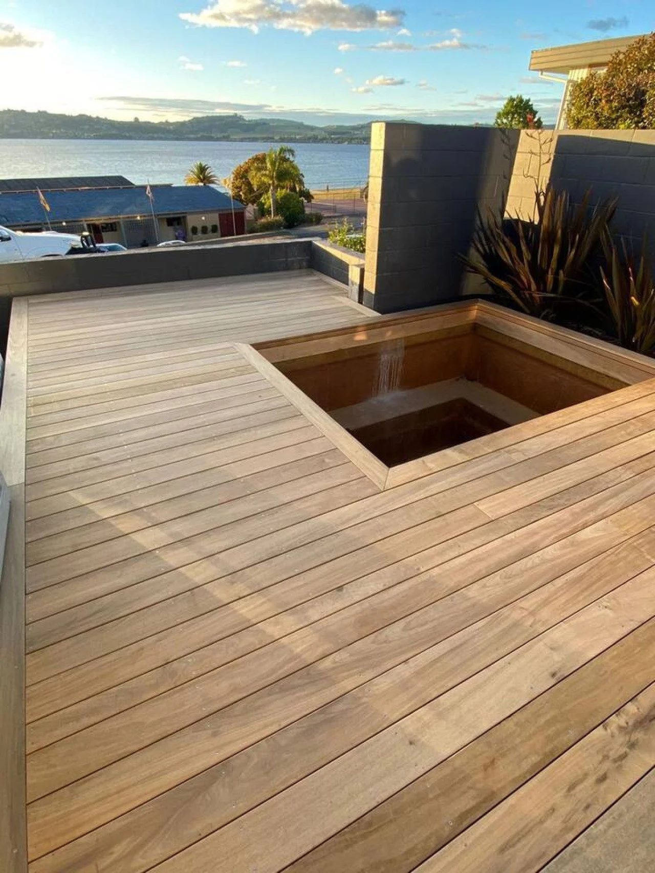 A wooden deck with a built-in hot tub overlooking a body of water, with trees and hills in the distance, and a partly cloudy sky.
