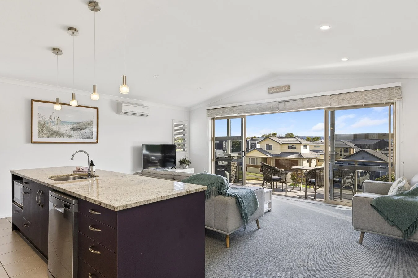 Living room with large sliding glass doors opening to a balcony with outdoor seating, a television, and a kitchen island with a sink and drawers.
