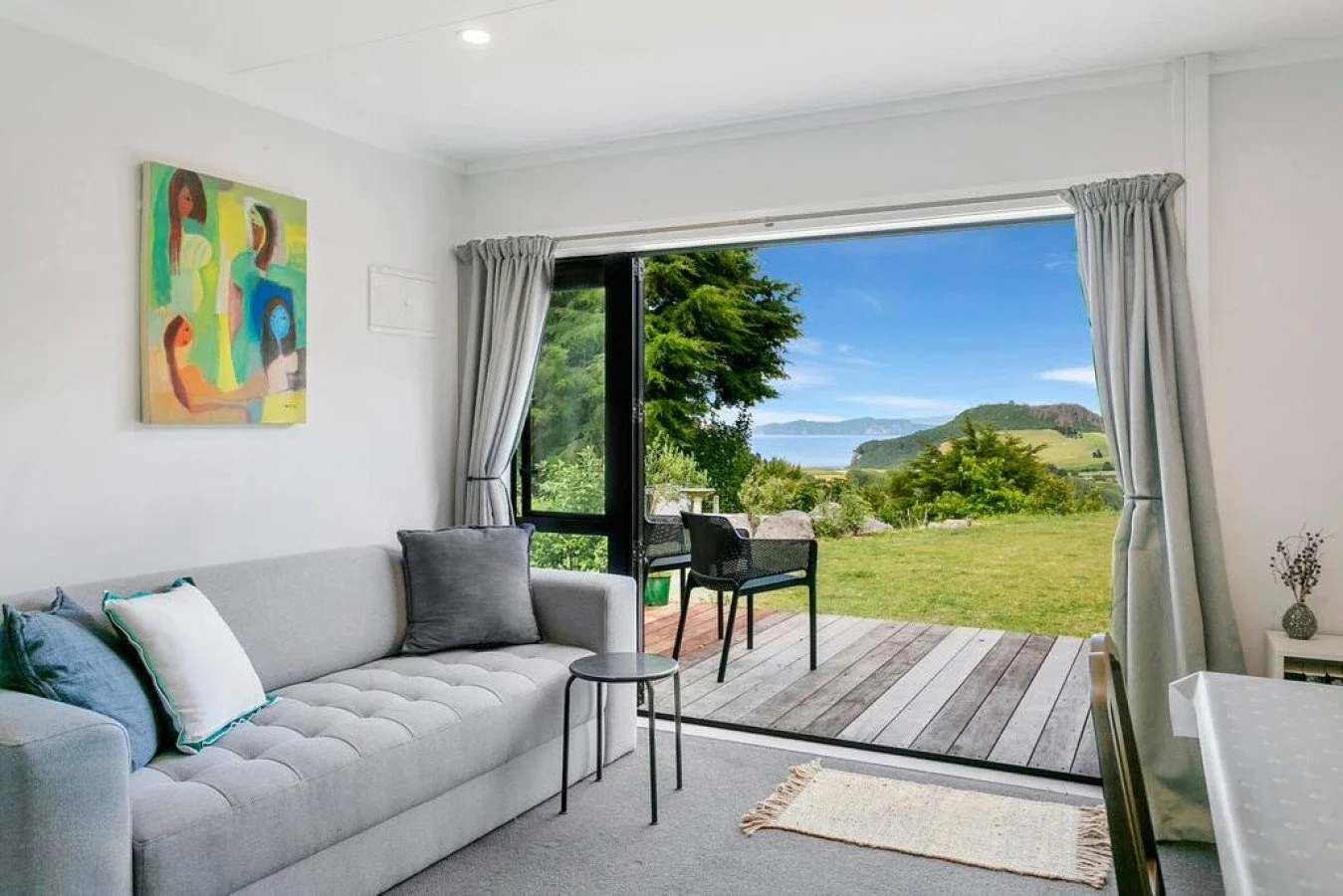 Living room with a gray sofa, decorative pillows, a small side table, artwork on the wall, and an open sliding door leading to a deck with outdoor chairs, overlooking a lush green landscape with trees, grass, and a body of water under a blue sky.