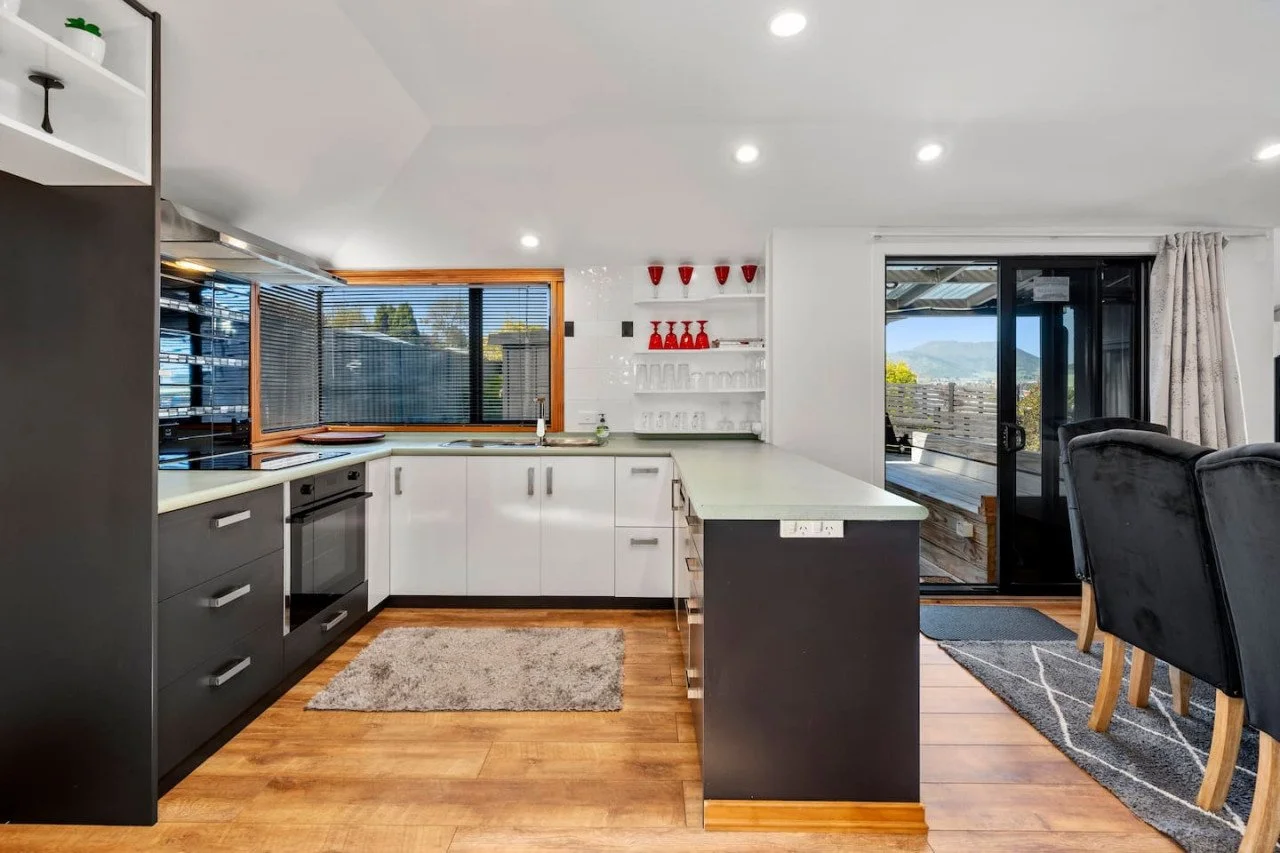 Modern kitchen with white and black cabinets, wooden floor, large window, open to a dining area with black chairs and a sliding glass door leading outside.