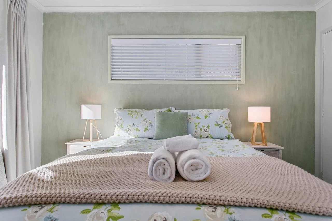 A neatly made bedroom with a bed featuring floral bedding and a green accent pillow, flanked by white nightstands with small lamps. Three rolled white towels are placed on the bed. There is a window with blinds behind the bed and cream curtains on th