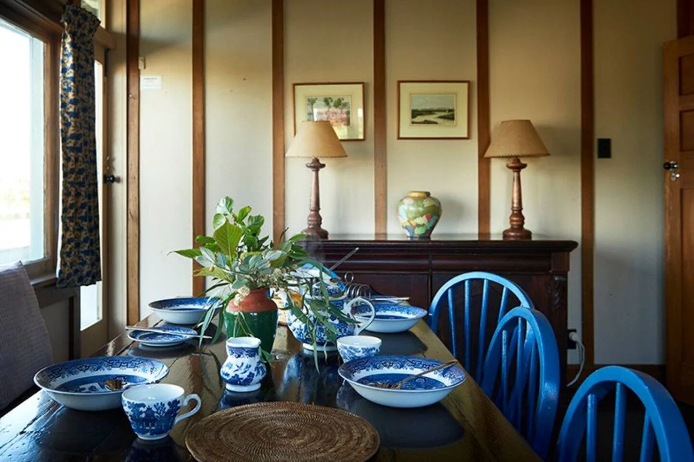 A dining room with a dark wooden table set with blue and white china, green plants, and wicker placemats. There are blue chairs around the table, beige lamps on a wooden sideboard, framed pictures on the wall, and a window with floral curtains.