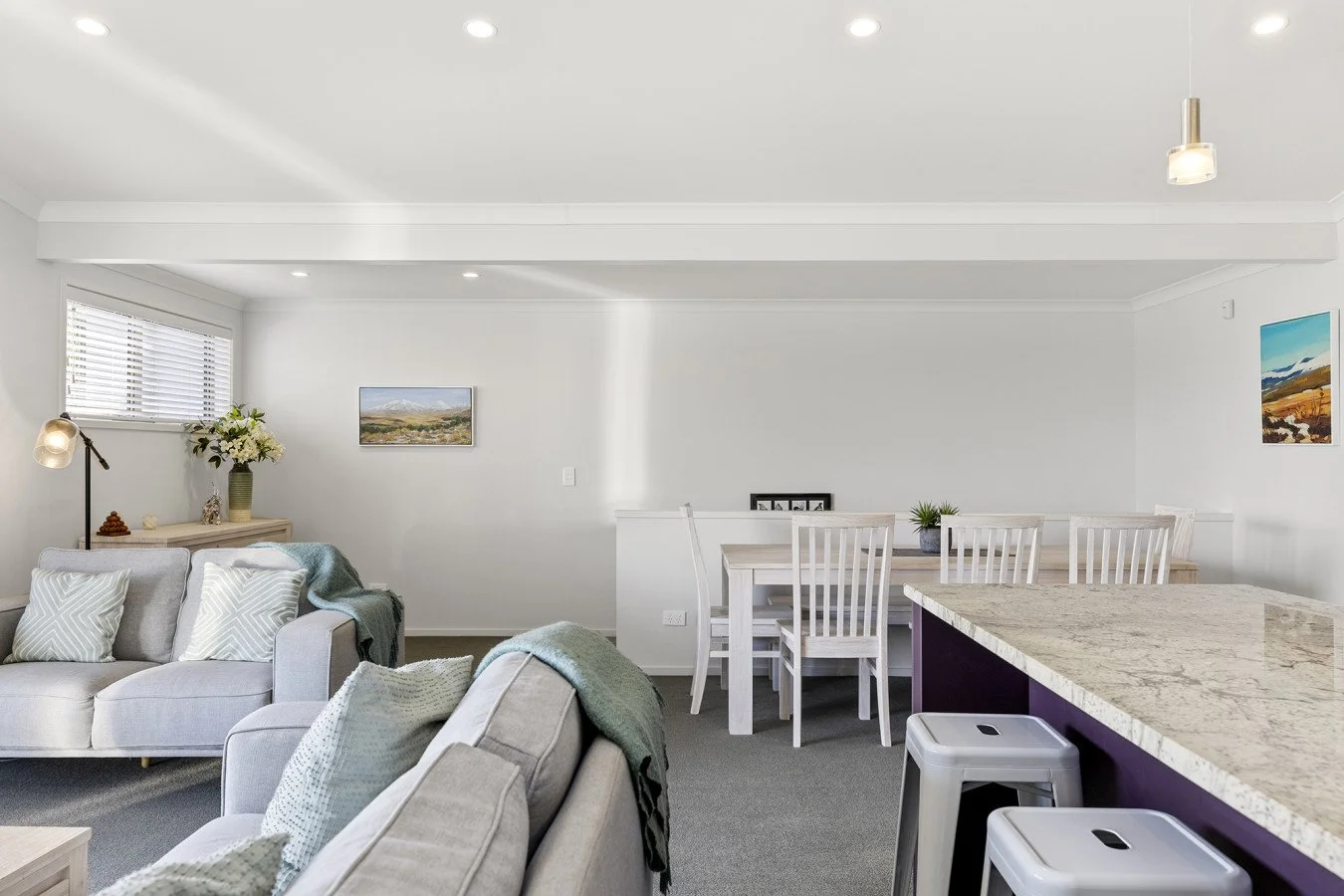 Modern living room and dining area with white walls, gray sofas with patterned pillows, a marble kitchen island with two barstools, wall art, a window with blinds, a hanging light, lamp with flowers, and minimalist decor.