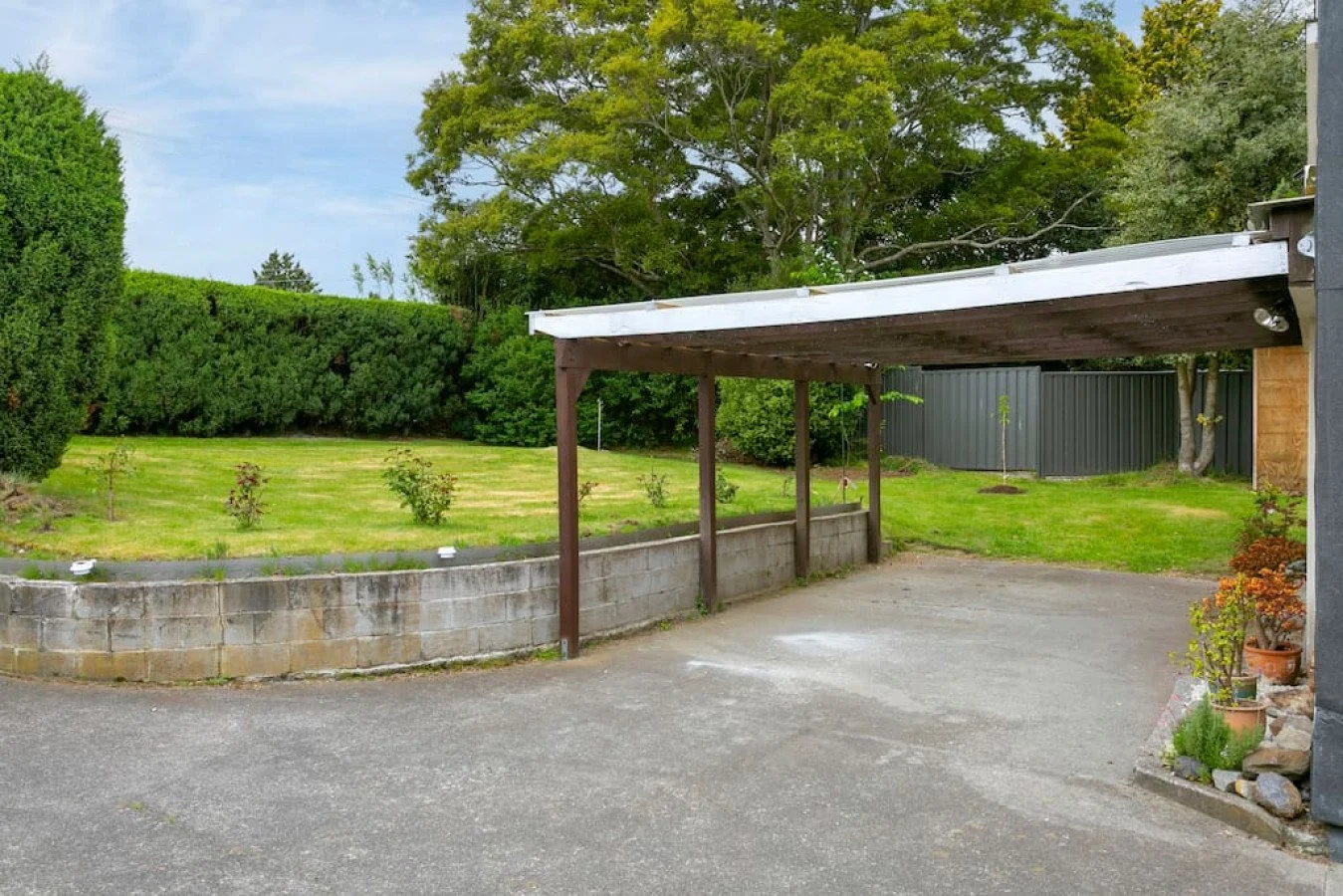 A backyard with a concrete driveway, a wooden carport, a grassy area with small bushes, tall trees, and a dark gray fence in the background.