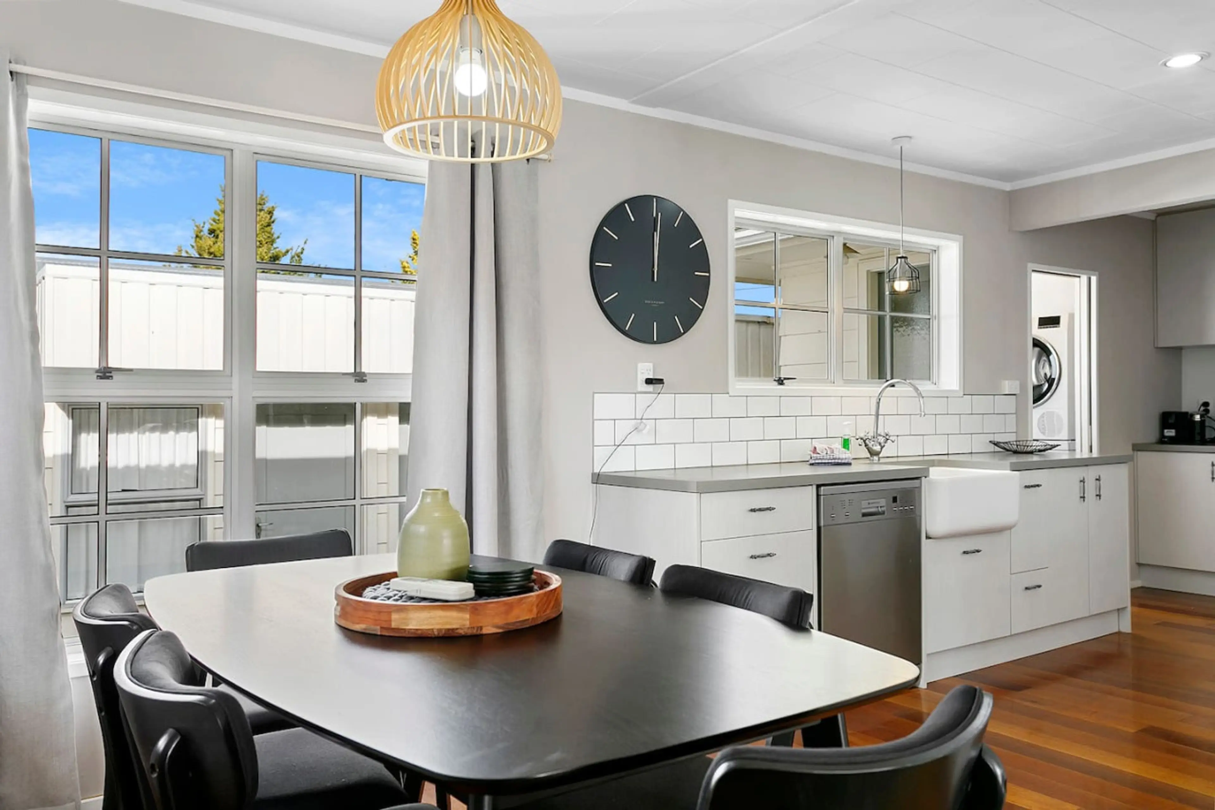 Modern kitchen and dining area with a black dining table and six black chairs, large windows with curtains, a wall clock, white tiled backsplash, and a dishwasher.