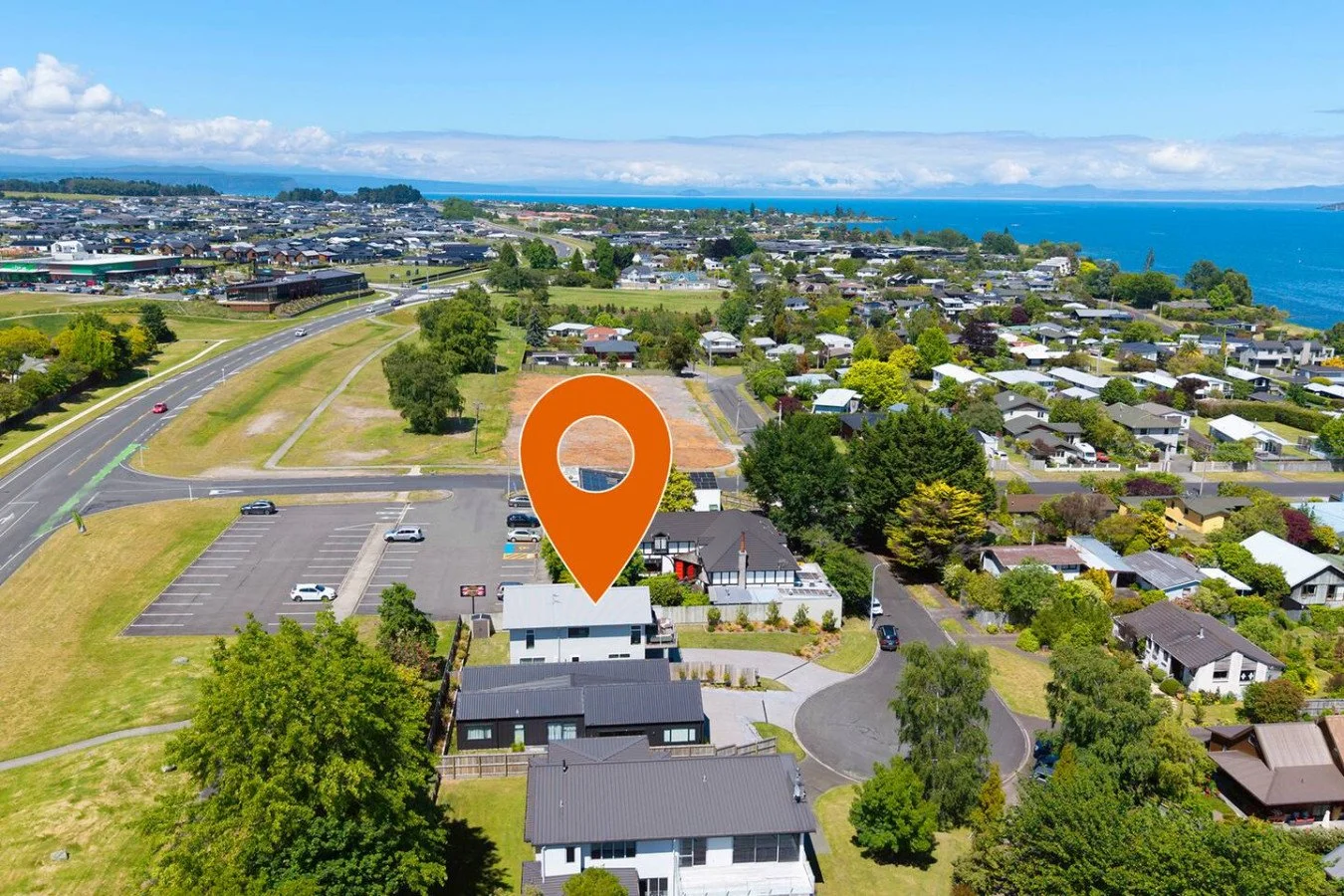 Aerial view of a suburban neighborhood with a coastline in the background, and a large orange map pin marker pointing to a specific house along the street.