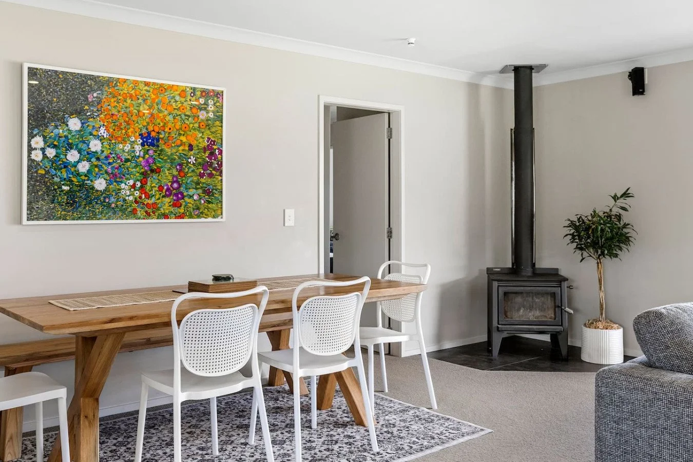 Living room with a wooden dining table, white chairs, a colorful floral painting on the wall, a black wood stove, a potted plant, and a section of gray sofa.