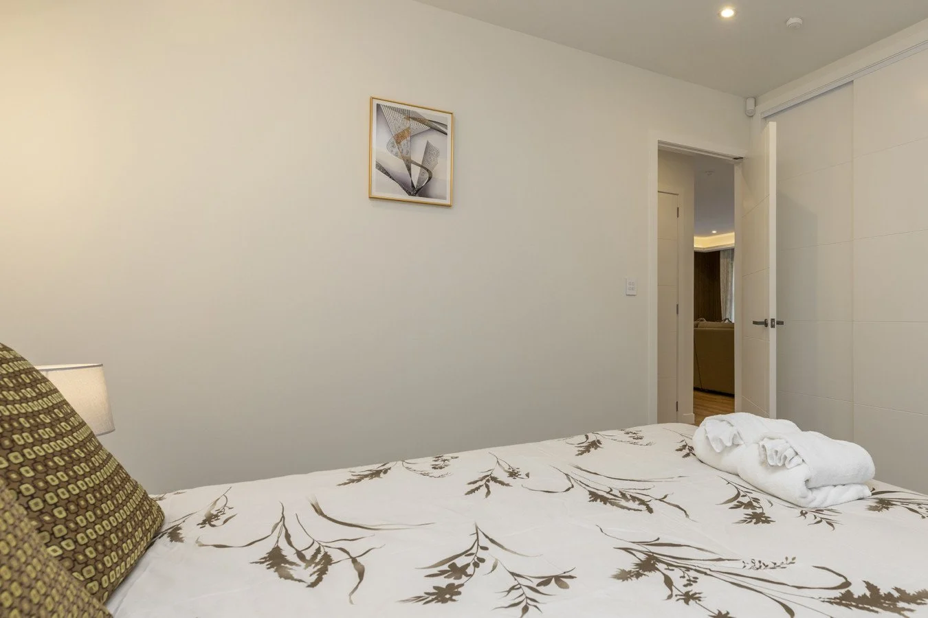 A neatly made bed with floral bedding and rolled white towels, in a modern hotel room with white walls and a small framed abstract art piece.