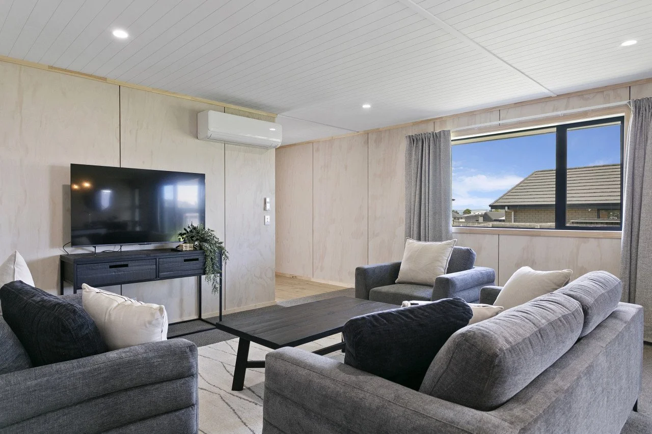 Living room with gray sofas, white pillows, a dark wood coffee table, a large flat-screen TV on a dark console, a window with gray curtains, and a view of a house roof outside.