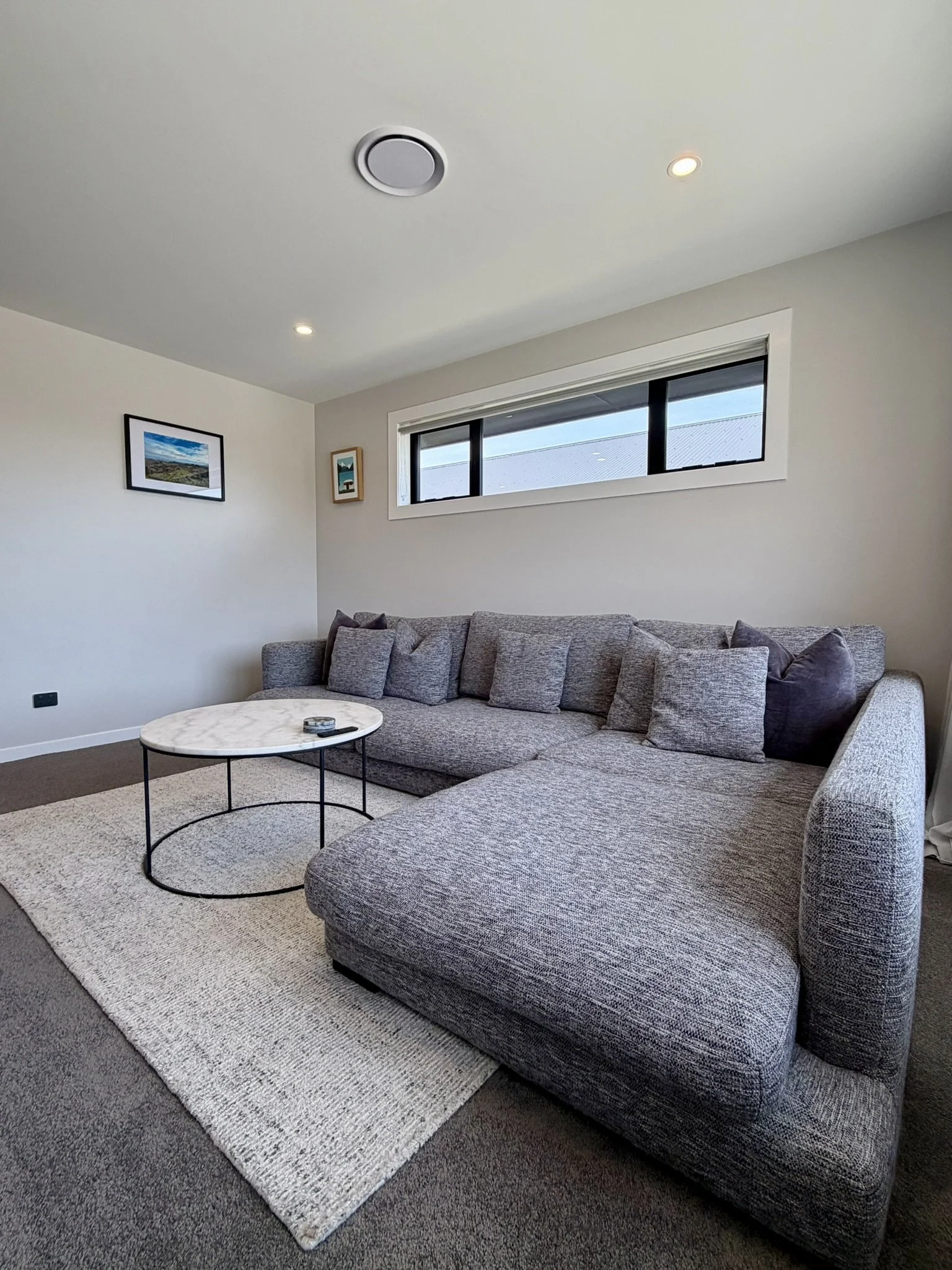 Living room with a gray L-shaped sectional sofa, a round marble-top coffee table, a white textured area rug, and three small framed pictures on the white wall. There is a horizontal window near the ceiling, and the room has white walls, a beige ceili