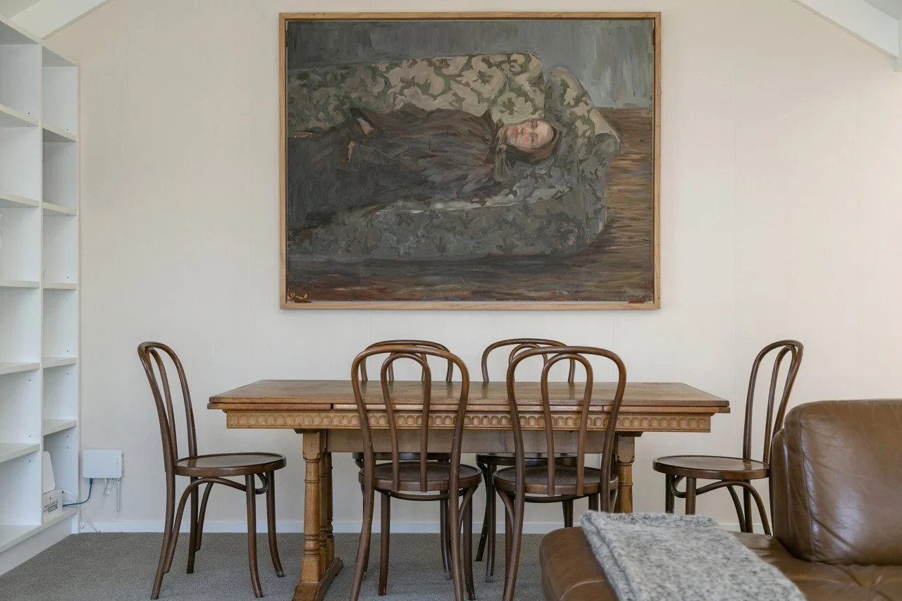 A dining room features a wooden table with six matching chairs, a large framed painting of a woman lying on a sofa, and an empty white bookshelf on the left. A leather armchair with a blanket and a gray carpet are partially visible.