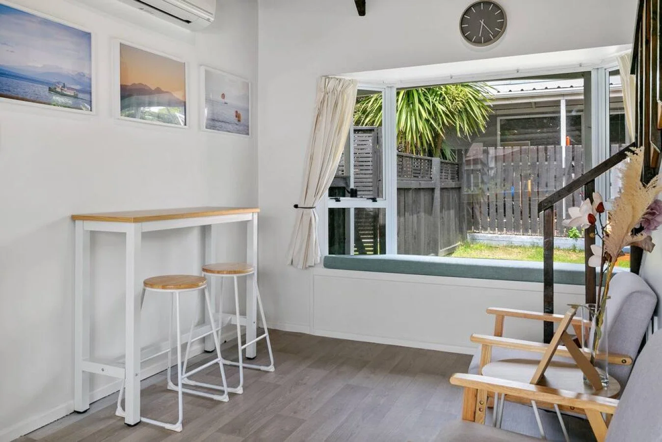 A cozy room with a window seat, potted plant, two bar stools at a high table, and wall art depicting beaches and mountains.