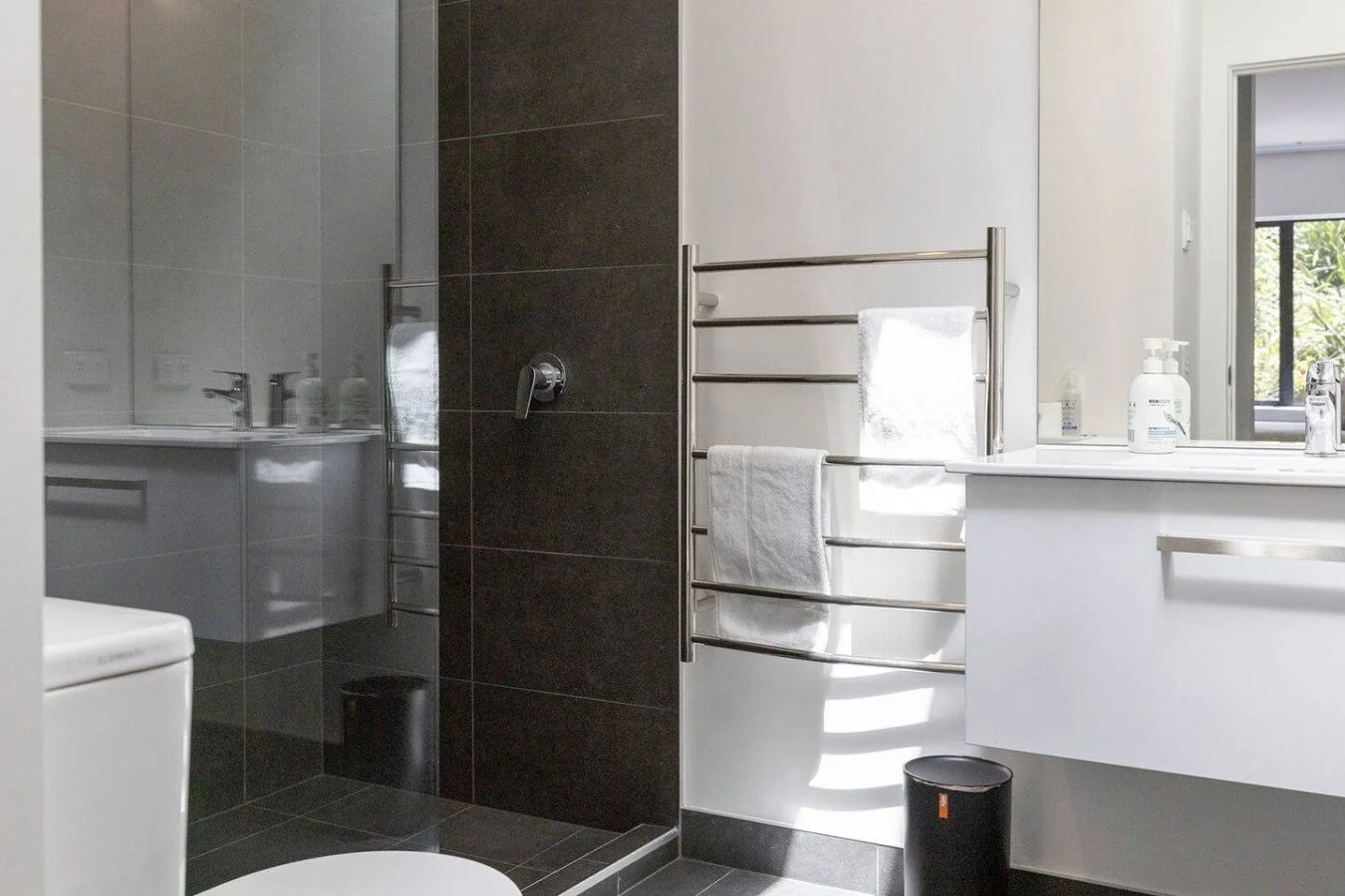 Modern bathroom with black and white tiles, a walk-in shower with a glass door, a towel rack with towels, a sink with toiletries, and a window with natural light.