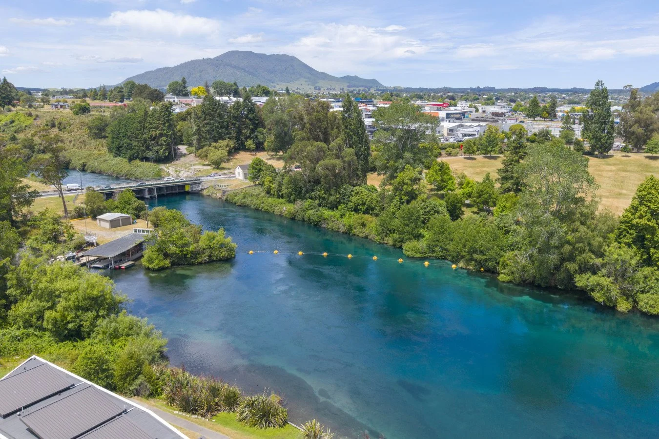 A scenic aerial view of a river with clear blue water flowing through a green landscape, surrounded by trees and grass, with a bridge crossing over the river and a shopping center visible in the background, under a partly cloudy sky.