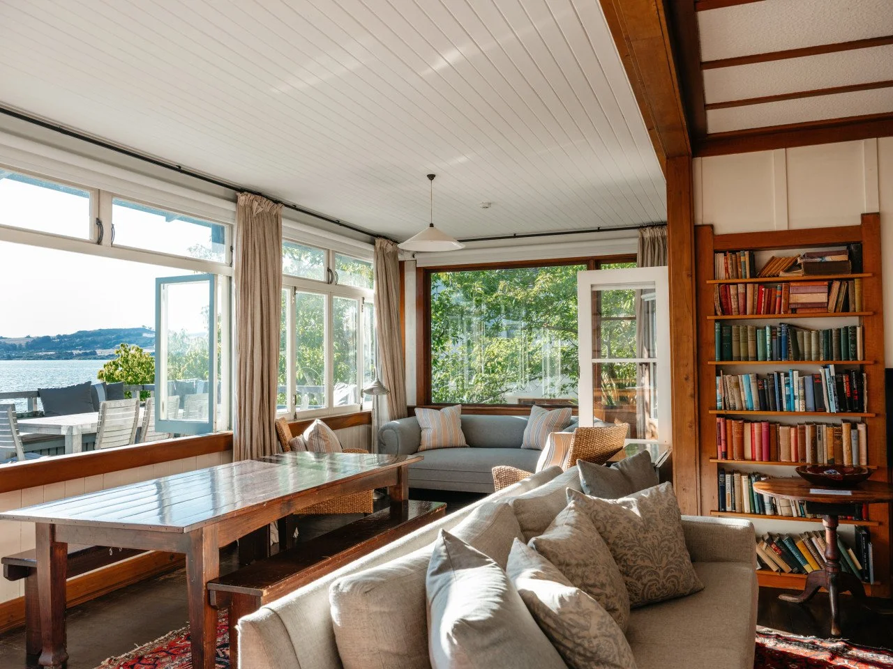 Living room with large windows revealing a lake view, beige sofa with patterned pillows, wooden bookshelf filled with books, dining table, and outdoor patio visible through open windows.