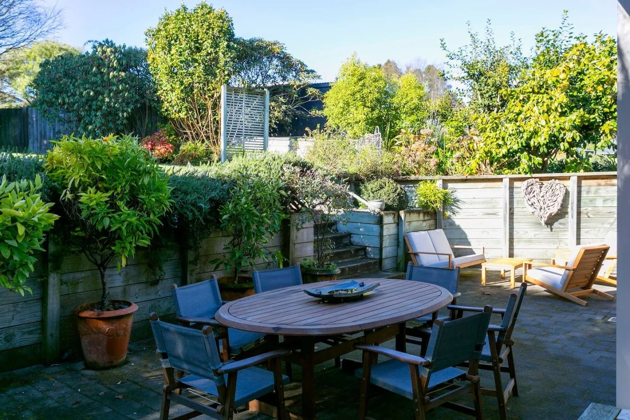 Outdoor patio with wooden table and six chairs, surrounded by potted trees, bushes, and a wooden privacy fence decorated with a heart-shaped ornament.