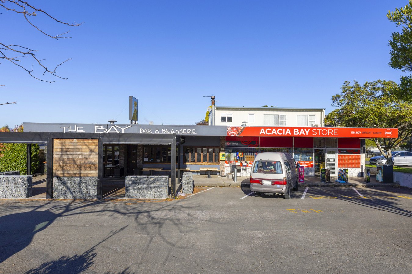 A small parking lot in front of a restaurant called The Bay Bar & Brasserie, with a red and white Acacia Bay Store next to it. There is a silver minivan parked in front of the store, and a few other cars visible on the street. The sky is clear and bl