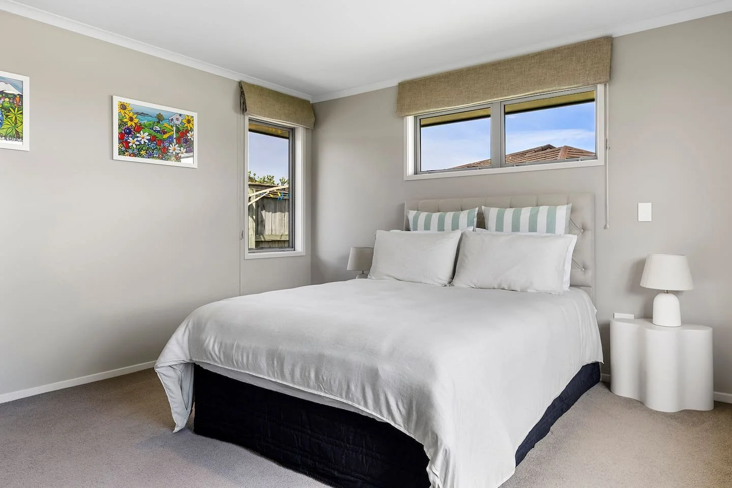 A neatly made bed with white sheets and pillows in a minimalistic bedroom, with two small windows and artwork on the wall.