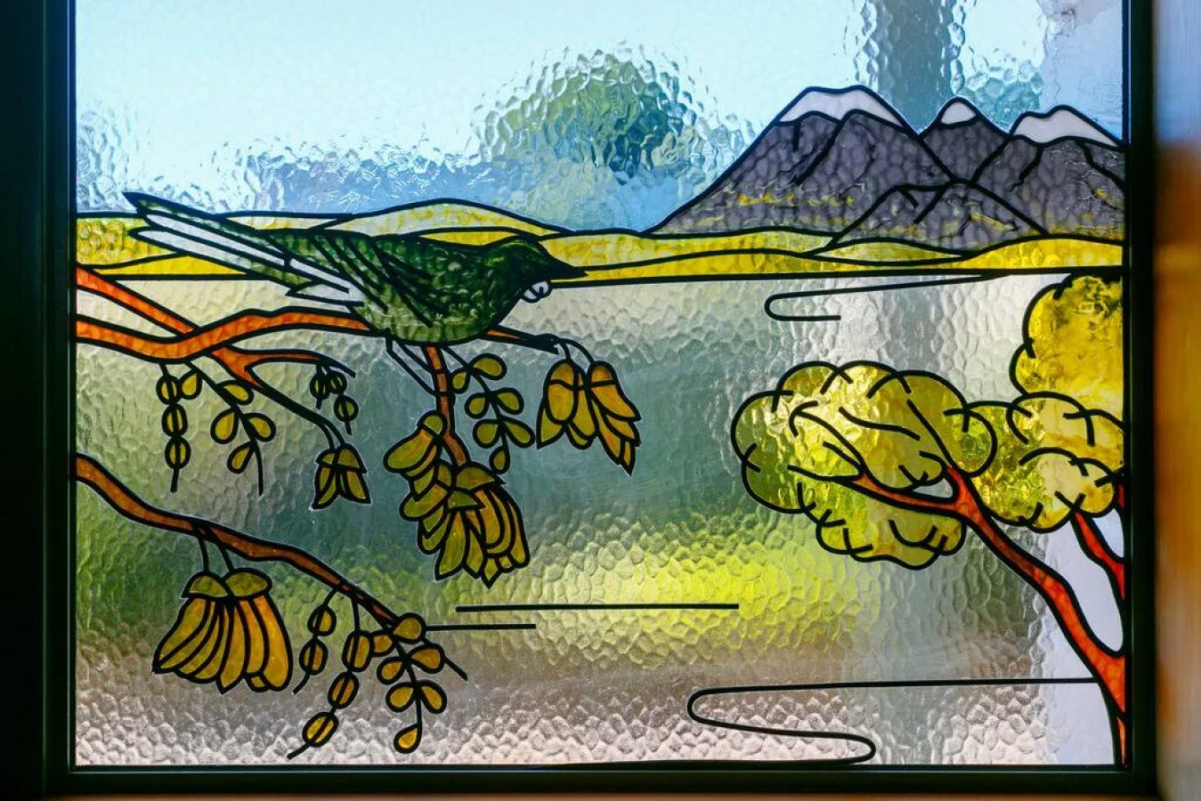 Colored stained glass window depicting a green bird perched on a branch with yellow leaves, mountains, and a lake or river in the background.